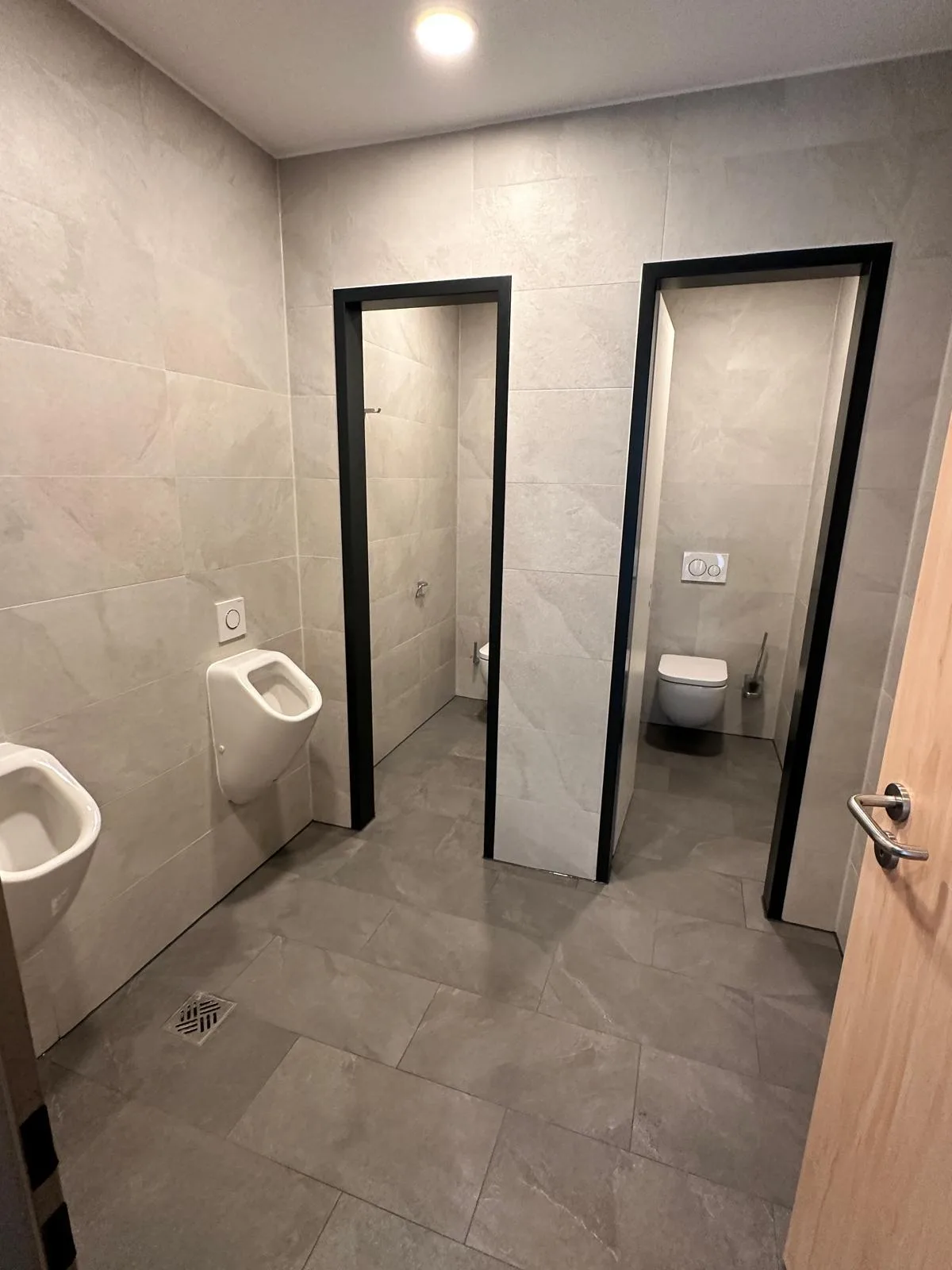 Geflieste Sanitäranlage mit Urinalen und separaten WC-Kabinen in grauer Steinoptik